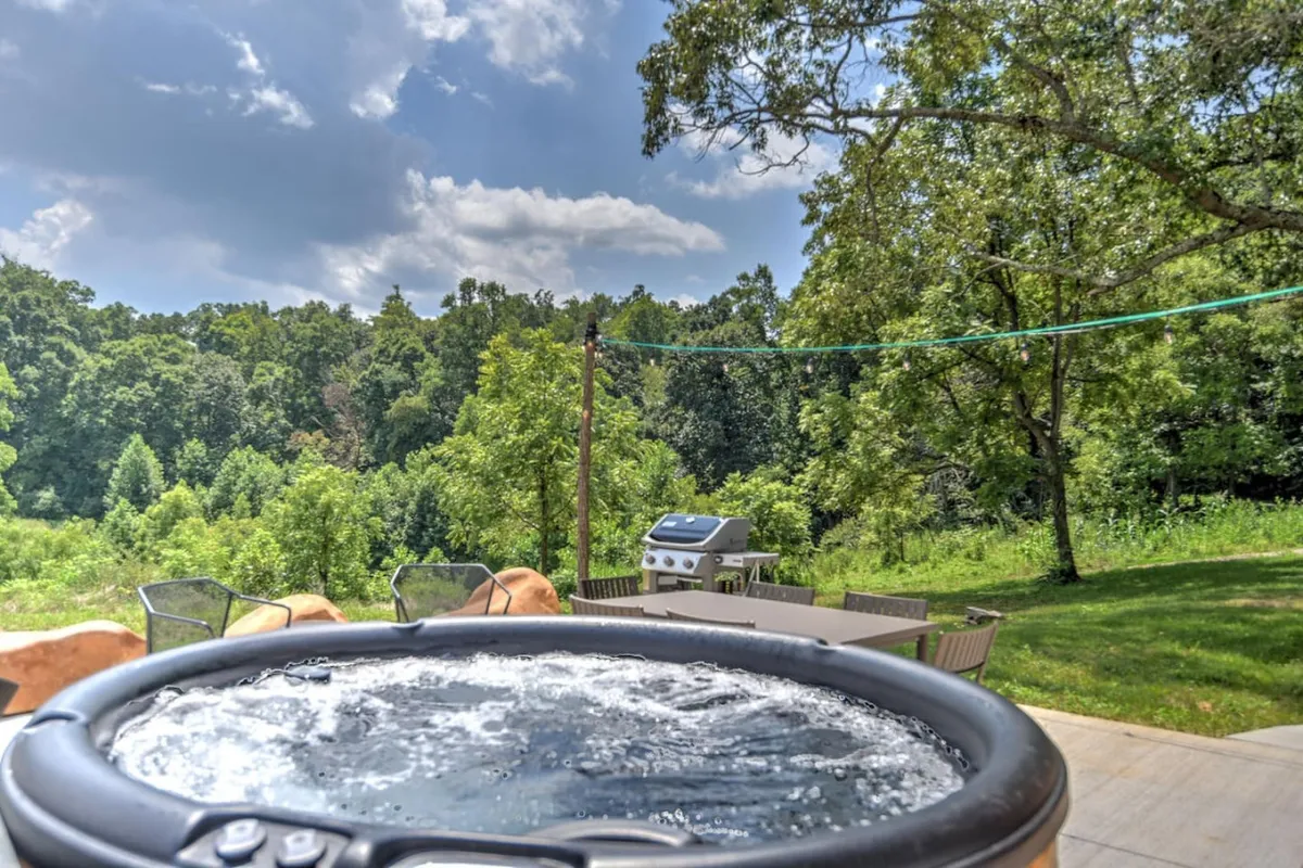 A spacious patio area showcasing a hot tub, grill, patio furniture, and abundant lush greenery. The area is inviting for outdoor relaxation, dining, or enjoying morning coffee with views of the natural landscape.
