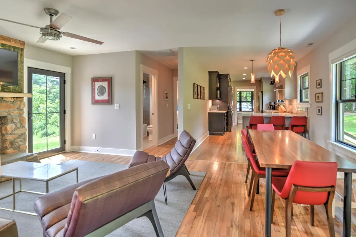 A wide view of the open-concept living, dining, and kitchen spaces with a visible stone fireplace, large windows, a dining table, and a modern kitchen in the background. The arrangement fosters easy movement, togetherness, and sociability.