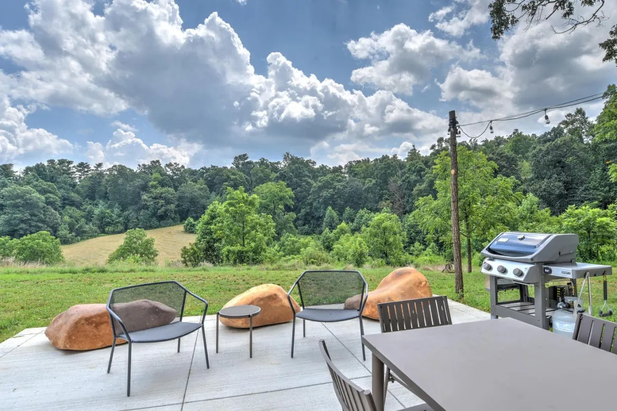 An exterior view featuring a grill, outdoor seating, large boulders, and a sweeping green landscape. The scene sets a welcoming tone for sunset views, social gatherings, and peaceful evenings under the open sky.