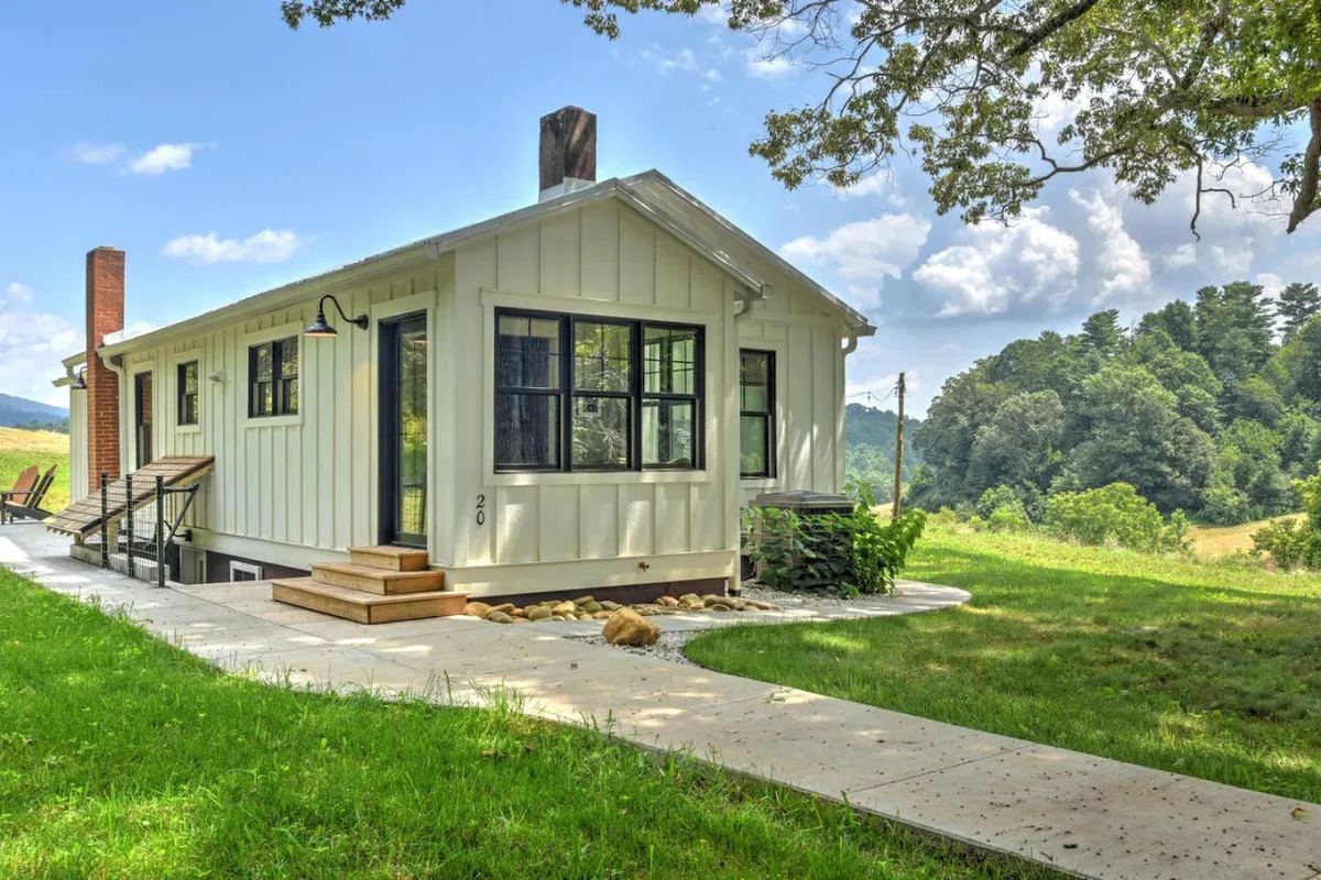 The image shows a charming farmhouse set amidst lush greenery, with a welcoming front porch, a winding pathway leading to the house, and mature trees surrounding the property. The peaceful setting hints at ample outdoor space perfect for relaxation, with the mountains of Western North Carolina in the background.