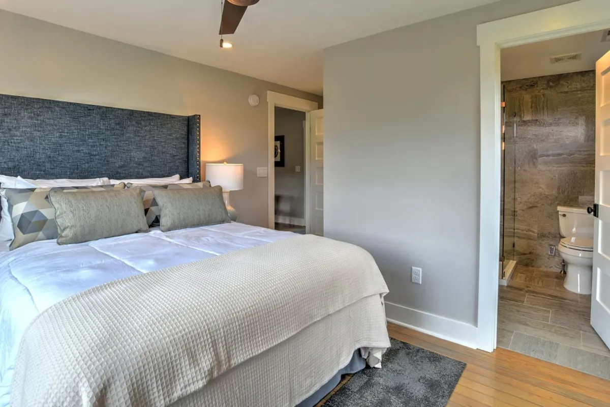 A peaceful bedroom scene featuring a bed adorned with decorative pillows, a softly lit bedside lamp, and an open bathroom door visible in the background. The design showcases plush bedding and cozy farmhouse-inspired details, evoking a sense of tranquility and comfort.