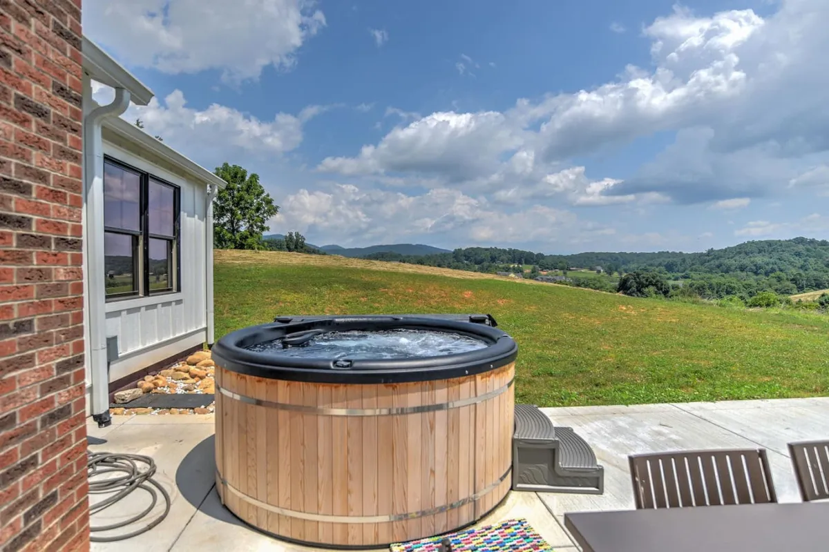 This image features a wooden hot tub set against a backdrop of expansive green landscape and sweeping mountain views. The patio area is spacious and inviting, with ample room for gatherings and outdoor meals. The tranquil atmosphere is highlighted by the serene setting and the natural beauty surrounding the hot tub.