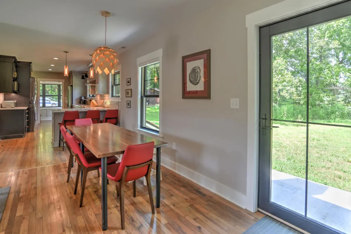 A spacious dining area anchored by a large wooden table surrounded by red chairs. Expansive windows provide views of the outdoors and flood the room with natural light, while artwork and homey touches foster a welcoming environment.