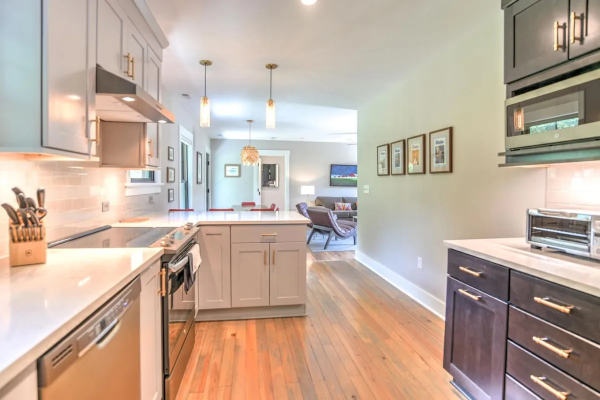 A bright, welcoming kitchen featuring a large island at center, complemented by stainless steel appliances, wooden flooring, rustic cabinetry, and modern hanging light fixtures. The space receives ample natural light, highlighting the farmhouse charm and fully equipped layout.