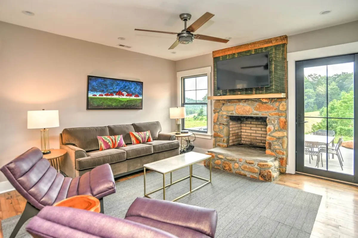 A cozy living room with a prominent stone fireplace (used for display only), exposed beams overhead, large windows letting in natural light, colorful artwork on the wall, and comfortable modern seating arranged to encourage conversation and relaxation.