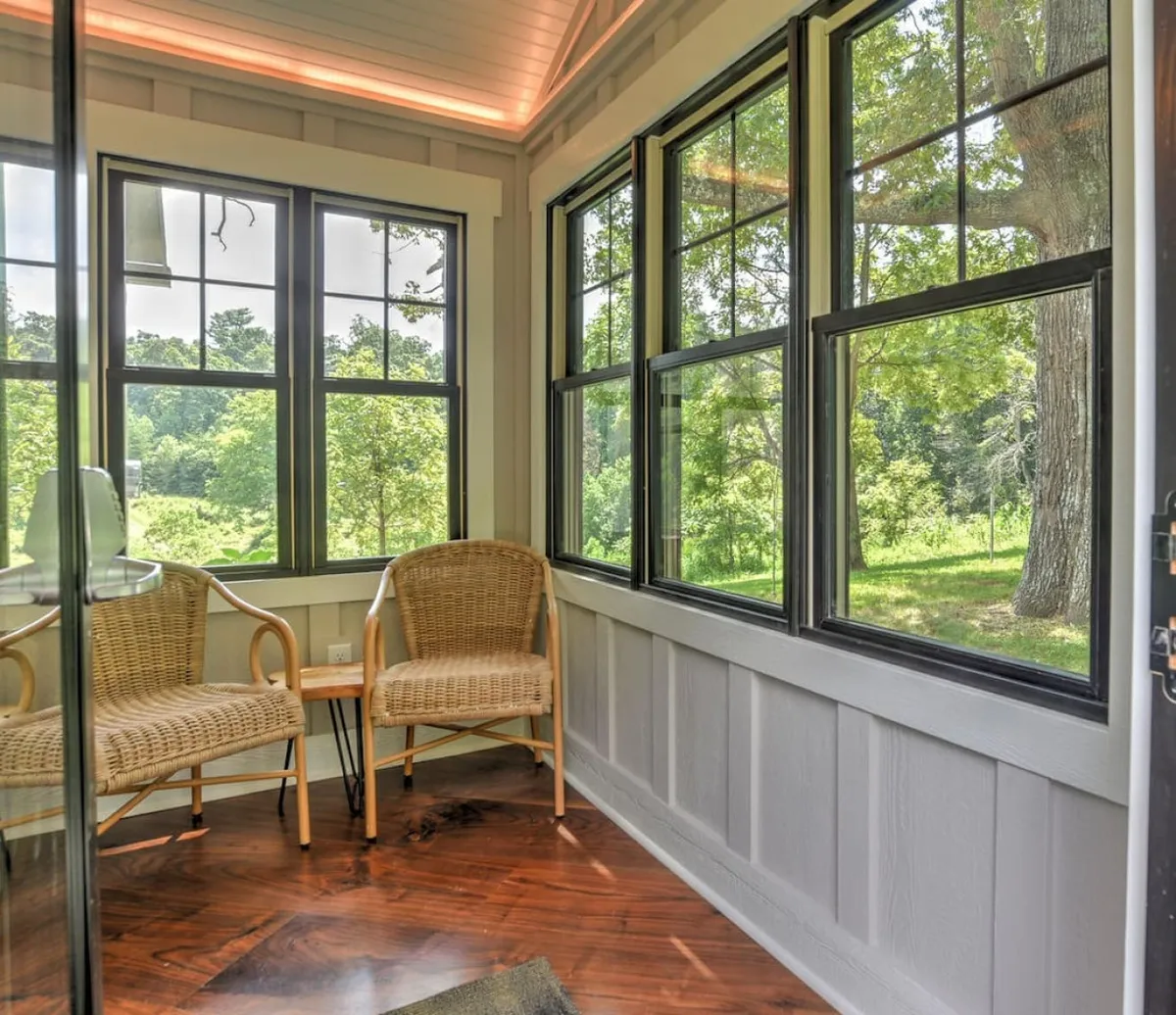 A serene interior nook featuring two wicker chairs and a small wooden table set by expansive windows, offering tranquil views of the surrounding landscape. The natural light and sightlines to the outdoors evoke a sense of connection to nature and a retreat-like atmosphere.