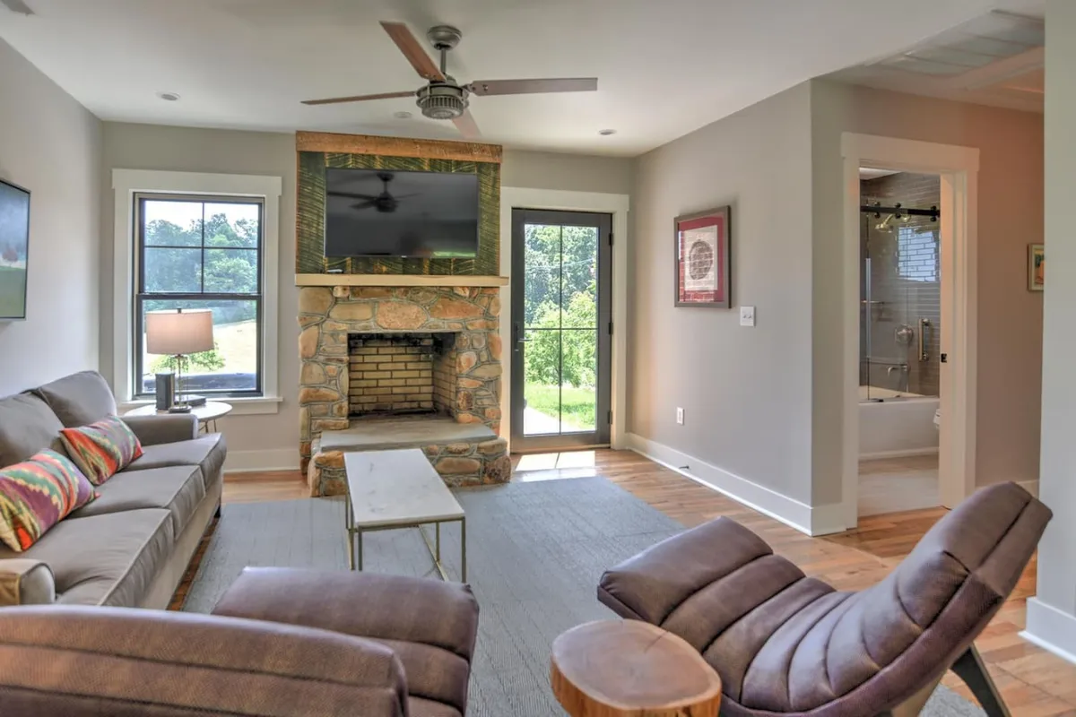 A welcoming living room with a stone fireplace (decorative), large windows flooding the space with natural light, comfortable seating areas, and a sliding glass door suggesting access to the outdoors. The room is designed for relaxation and family gatherings, with views connecting the interior to the surrounding landscape.